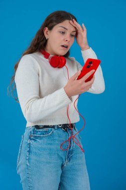 Beyaz kadın akıllı telefonundan kötü haberler okurken şok olmuş görünüyor. Kulaklık takıyor ve beyaz bir kazak giyiyor. Katı mavi arka plana karşı kaygıyla tepki veriyor..