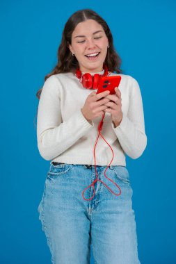 Kırmızı kulaklığıyla ayakta duran ve müziği seven gülümseyen genç bir kadın kırmızı bir akıllı telefon tutarken, beyaz bir kazak ve kot pantolon giyerken, dikey mavi bir arka plana karşı. 