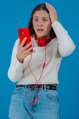 Beyaz kadın akıllı telefonundan kötü haberler okurken şok olmuş görünüyor. Kulaklık takıyor ve beyaz bir kazak giyiyor. Katı mavi arka plana karşı kaygıyla tepki veriyor..