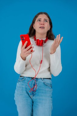 Beyaz kadın akıllı telefonundan kötü haberler okurken şok olmuş görünüyor. Kulaklık takıyor ve beyaz bir kazak giyiyor. Katı mavi arka plana karşı kaygıyla tepki veriyor..