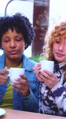 Kafe masasında oturan iki kadın, birlikte sıcak içecekler içerler, gündelik sohbet, dostluk, rahatlama anı, modern yaşam tarzı, gündüz vakti, kentsel ortam, gündelik moda, dikey imaj 