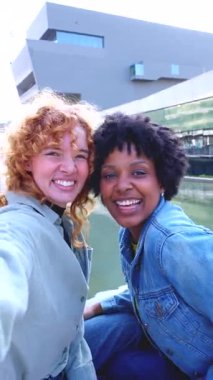 Dışarıda oturan mutlu, el sallayan ve gülümseyen genç kadınlar video çağrısı, dijital iletişim, arkadaşlık, kentsel geçmiş, neşeli duygular, gündelik moda, eğlenceli bağlantı. 