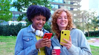 İki mutlu bayan arkadaş, akıllı telefonlar kullanarak, sosyal medya uygulamalarını kontrol ederek, şehir parkında oturarak, gülümseyerek, arkadaşlıkla, modern teknolojiyle, iletişimle, yaşam tarzıyla, 