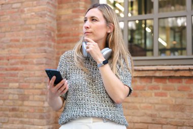 Genç bir kadın dışarıda duvara yaslanmış, boynuna kablosuz kulaklık takmış, tek eliyle akıllı bir telefon tutuyor, dalgın dalgın bakıyor.. 