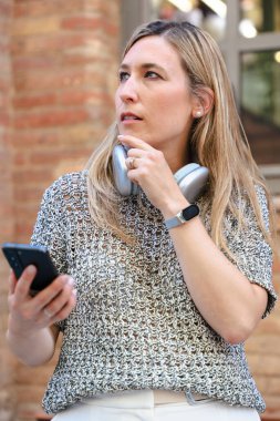 Genç bir kadın dışarıda duvara yaslanmış, boynuna kablosuz kulaklıklar takmış, tek eliyle akıllı bir telefonu sıkıca tutuyor, yandan eğilip dalgın dalgın bakıyor. Dikey
