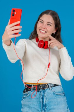 Kırmızı akıllı telefon ve kulaklıkla selfie çeken mutlu genç bir kadının dikey görüntüsü. Mavi bir arkaplan önünde, gündelik ve modern bir dijital anın tadını çıkararak güvenle gülümsüyor. Dikey