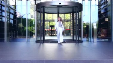 Beyaz takım elbiseli bir iş kadını, elinde akıllı bir telefon, profesyonel, kentsel bir şirket ortamı olan döner bir kapıdan modern cam ofis binasından çıktıktan sonra dışarıya doğru yürür. 
