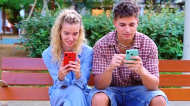 Genç bir erkek ve kadın parkta bir bankta oturuyor, her biri renkli bir akıllı telefon tutuyor, sosyal medya uygulamalarını kontrol ediyor ve paylaşımları karşılaştırırken gülümsüyor, günlük yaz kıyafetleri, arkadaşça bir an. 