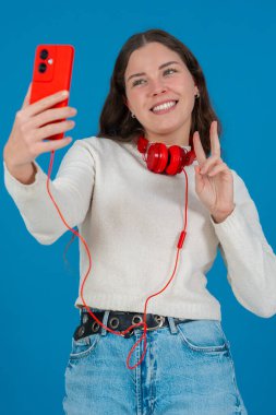 Kırmızı akıllı telefon ve kulaklıkla selfie çeken mutlu genç bir kadının dikey görüntüsü. Mavi bir arkaplan önünde, gündelik ve modern bir dijital anın tadını çıkararak güvenle gülümsüyor. Dikey