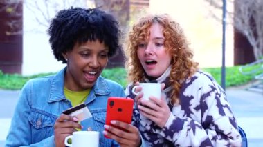 Dışarıda oturan neşeli genç kadınlar, birlikte kahvenin tadını çıkarırken bir yandan da kredi kartıyla internet üzerinden alışveriş yapıyorlar. Her iki arkadaş da gülümser ve güneşli bir günde neşeli bir ortam paylaşır.. 