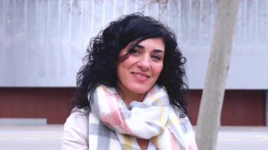 Kıvırcık siyah saçlı, atkı ve palto giyen kendine güvenen beyaz kadın şehir ortamında dışarıda dikiliyor. Modern yaşam tarzı, şehir modası ve kendinden emin ifade serin bir günde. 