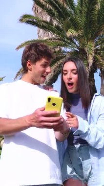 Güneşli bir parkta yeşil çimlerde birbirine yakın gülümseyen genç bir çift akıllı telefon kullanarak boş vakitlerin tadını çıkarıyor. Günlük kıyafetler, arka planda palmiye ağaçları, açık havada eğlenceli anlar..