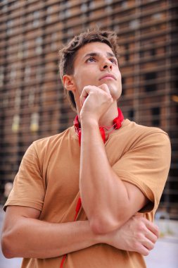 Boynuna kahverengi tişört ve kırmızı kulaklık takan dalgın genç bir adam modern ahşap bir duvarın önünde dikiliyor, eli çenesinde düşünceli bir şekilde yukarı bakıyor.