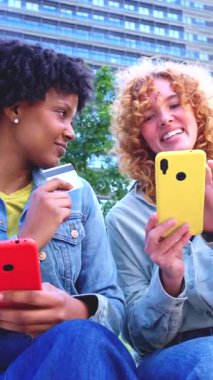 Şehir parkında oturmuş, akıllı telefonlar kullanarak gülümseyen neşeli genç kadınlar. Birinin elinde kırmızı telefon ve kredi kartı var, diğerinde ise internet alışverişini veya cep telefonu ödemesini temsil eden sarı bir telefon..