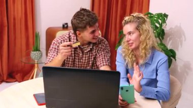 Bir masada oturan gülümseyen genç çift online alışveriş yapıyor, dizüstü bilgisayar, akıllı telefon ve kredi kartı kullanıyor. Kadın telefona bakarken adam kartı tutuyor, birlikte alışverişin tadını çıkarıyor.