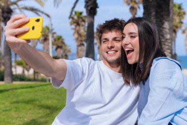 Genç bir adam ve kadın, güneşli bir avuç içinde bir bankta oturuyor. Deniz kenarında, gülümsüyor ve mutlu bir selfie çekerken yüzlerini çeviriyorlar. Akıllı telefonlar, somutlaştırıcı eğlence, arkadaşlık ve yaz titreşimleri.. 