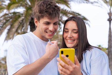 Güneşli bir parkta birlikte duran mutlu genç çift, sarı bir akıllı telefona bakarken gülümsüyor. Arka planda palmiye ağaçları, arkadaşlık ve teknoloji kullanımı olan rahat bir açık hava sahnesi 