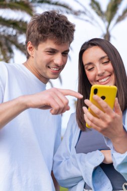 Güneşli bir parkta eğlenen mutlu genç çift, erkeğin kadının elinde tuttuğu akıllı telefon ekranını işaret ederken gülümsüyor. Palmiye ağaçları ve açık hava manzarası rahatlık yaratır. 