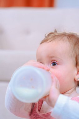 Halıda oturan ve sıcak bir evde şişeden su içen bir kızın yan görüntüsü. Yatay kompozisyon çocukluk, beslenme, büyüme ve erken bakım gibi konular için idealdir. Dikey