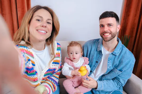 Mutlu, genç bir aile evde selfie çekiyor, anne, baba ve bebek birlikte kanepede oturuyor, keyifli ebeveynlik ve bağlanma anlarını yakalıyorlar rahat bir oturma odasında.