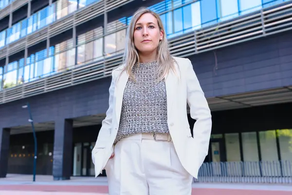 Hafif bir takım elbise giyen, ciddi şekilde kameraya bakan, modern bir ofis binasının önünde duran, liderliği, kararlılığı ve profesyonelliği temsil eden kendine güvenen bir iş kadını.. 