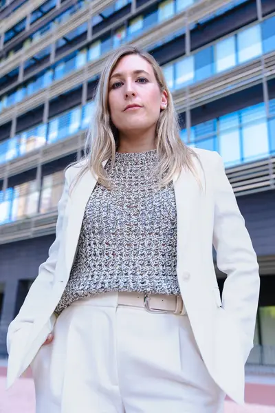Hafif bir takım elbise giyen, ciddi şekilde kameraya bakan, modern bir ofis binasının önünde duran, liderliği, kararlılığı ve profesyonelliği temsil eden kendine güvenen bir iş kadını.. 