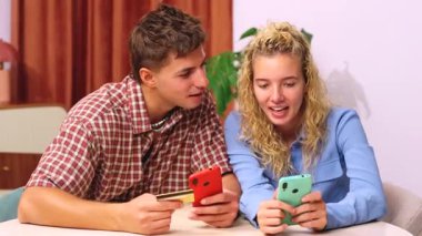 Bir masada oturan gülümseyen genç çift online alışveriş yapıyor, dizüstü bilgisayar, akıllı telefon ve kredi kartı kullanıyor. Kadın telefona bakarken adam kartı tutuyor, birlikte alışverişin tadını çıkarıyor.