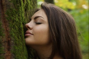 Genç bir kadın ağaç gövdesine dokunuyor, ruhsal iyileşme, ekolojik doğaya bağlanıyor. Doğanın tadını çıkar. Ağaçta yosun var. Yeşil bir ormanda temiz hava soluyan rahat bir kadının profili. Yüksek kaliteli fotoğraf.