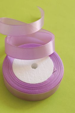 Bir rulo leylak saten kurdele canlı yeşil bir arka planda zarif bir şekilde kıvrılır. Yumuşak dokusu ve yumuşak pastel rengi. El işleri, hediye paketleme ve dekoratif projeler için mükemmel. Yüksek kalite fotoğraf
