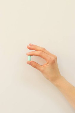 Narin bir el, yumuşak ve tarafsız bir arkaplana karşı küçük yeşil bir hap tutar. Minimalist kompozisyon, temiz ve modern bir estetik yaratarak sağlık, tıp ve refahı vurguluyor..