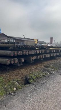 Bulutlu bir günde endüstriyel bir binanın yanında depolanmış, beton desteklere yerleştirilmiş, kırsal altyapı projelerine yerleştirilmeye hazır ahşap elektrik direkleri yığını. Yüksek kalite 4k görüntü