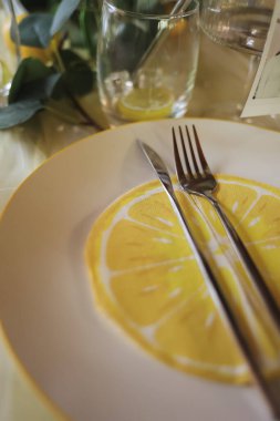  Limon temalı bir tabak, paslanmaz çelik çatal bıçak ve arka planda bir bardak ile zarif bir yaz sofrası. Taze ve canlı bir dekor, bir bahçe partisi ya da turunçgillerden esinlenilmiş bir kutlama için mükemmel.. 