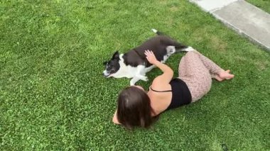  Genç bir kadın yemyeşil çimenlerin üzerinde uzanır siyah beyaz bir köpekle, nazikçe okşar ve bağlanır. Rahatlatıcı, sevgi, güven ve açık havada evcil hayvan ve insan arasında arkadaşlık anı. 4k görüntü