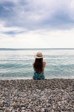  Hasır şapkalı bir kadın çakıl taşı plajında tek başına oturmuş bulutlu bir gökyüzünün altında denize bakıyor. Huzurlu seyahat anı yalnızlığı, özgürlüğü ve okyanusun farkındalığını simgeliyor. Yüksek kalite fotoğraf