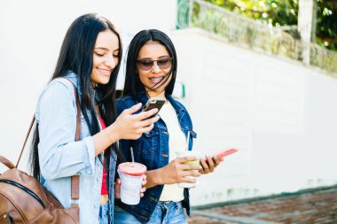 İki güzel ve mutlu İspanyol kadın sokakta gülümsüyor kontrol ediyor ve telefonları paylaşıyor. teknoloji kavramı.