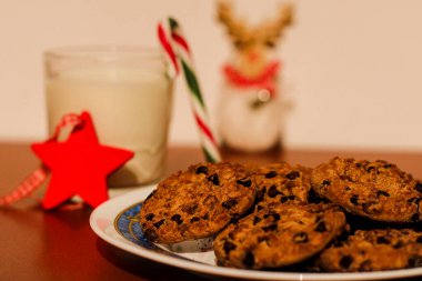 Ön planda bir bardak süt, Noel süsü, şeker kamışı ve beyaz arka planla bir tabak çikolatalı kurabiye..