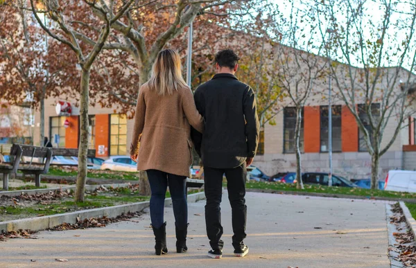 A young white couple walking backwards in a city on a park path, with trees in the background, arm in arm.