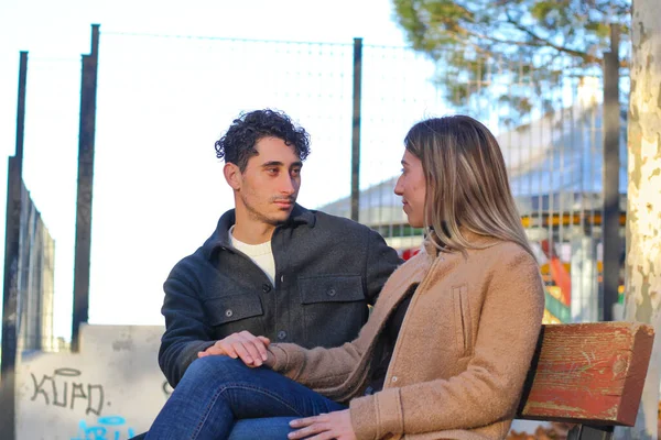 A young white couple sitting on a bench, looking at each other and holding hands.