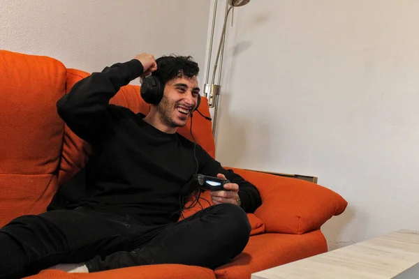 Single young white guy playing video game console with curly hair, on the sofa inside the house, excitedly, with large headphones
