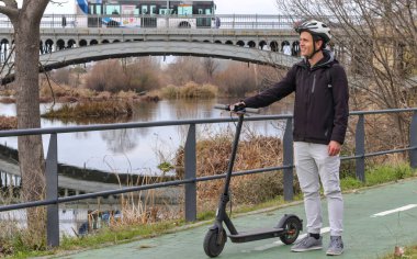 Elinde elektrikli scooter olan genç beyaz bir adam, miğfer ve sırt çantasıyla arka planda nehir ve köprü ile belirlenmiş bir şeritte duruyor..