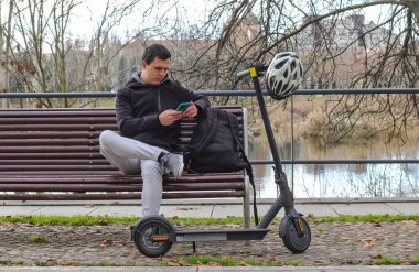 Genç beyaz adam elektrikli scooterıyla bankta oturuyor, cep telefonu kullanıyor, sırt çantası ve kaskı var, arka planda şehir var, denekler ortada..