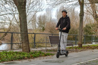 Genç beyaz adam elektrikli scooter kullanıyor, belli bir şeritte, kasklı ve sırt çantalı, doğal bir bölgede..