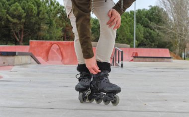 Arka plandaki kaykay parkında paten kayan genç beyaz bir adam. Yarı uzunlukta, ayakta, çömelmiş, arka planda eşofman altı ve ağaçlarla tanınmaz halde.