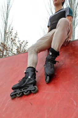 Siyah kaykaylı genç beyaz adam bir kaykay parkının rampasında oturuyor, antrenmandan sonra dinleniyor, sıradan elbiseler giyiyor, aşağıdan bakıyor, geleceğe bakıyor.