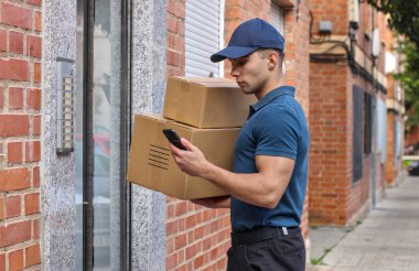 Üniformalı paket teslimatı yapan yakışıklı beyaz adam az önce geldi ve adresi kontrol etmek için cep telefonuna bakıyor. Çevrimiçi alışveriş kavramı