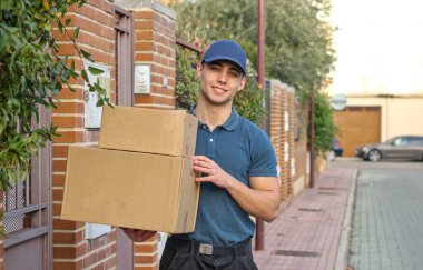 Yakışıklı, üniformalı, gülümseyen, sokakta durmuş elinde paketlerle kameraya bakan genç bir adam. Çevrimiçi alışveriş kavramı