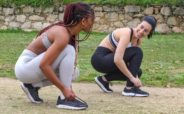 Young Caucasian woman and young black woman tying sneakers outdoor, looking at each other and smiling, getting ready to run, dressed in sportswear, side view. Concept of running