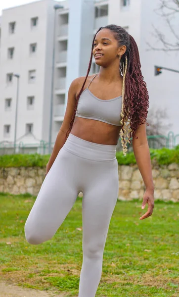 Young and pretty black woman stretching her leg outdoor in sportswear, getting ready to run, looking away, front view, vertical, building in the background. Concept of running