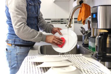 Bulaşıkları süngerle yıkayan bir adam. Yanında yığılmış bir adam. Tanınmaz halde. Ev işi yapan erkek kavramı