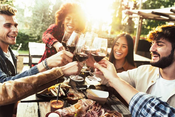 Multiracial people cheering red wine sitting outside at bar table. Social gathering, youth and beverage lifestyle concept
